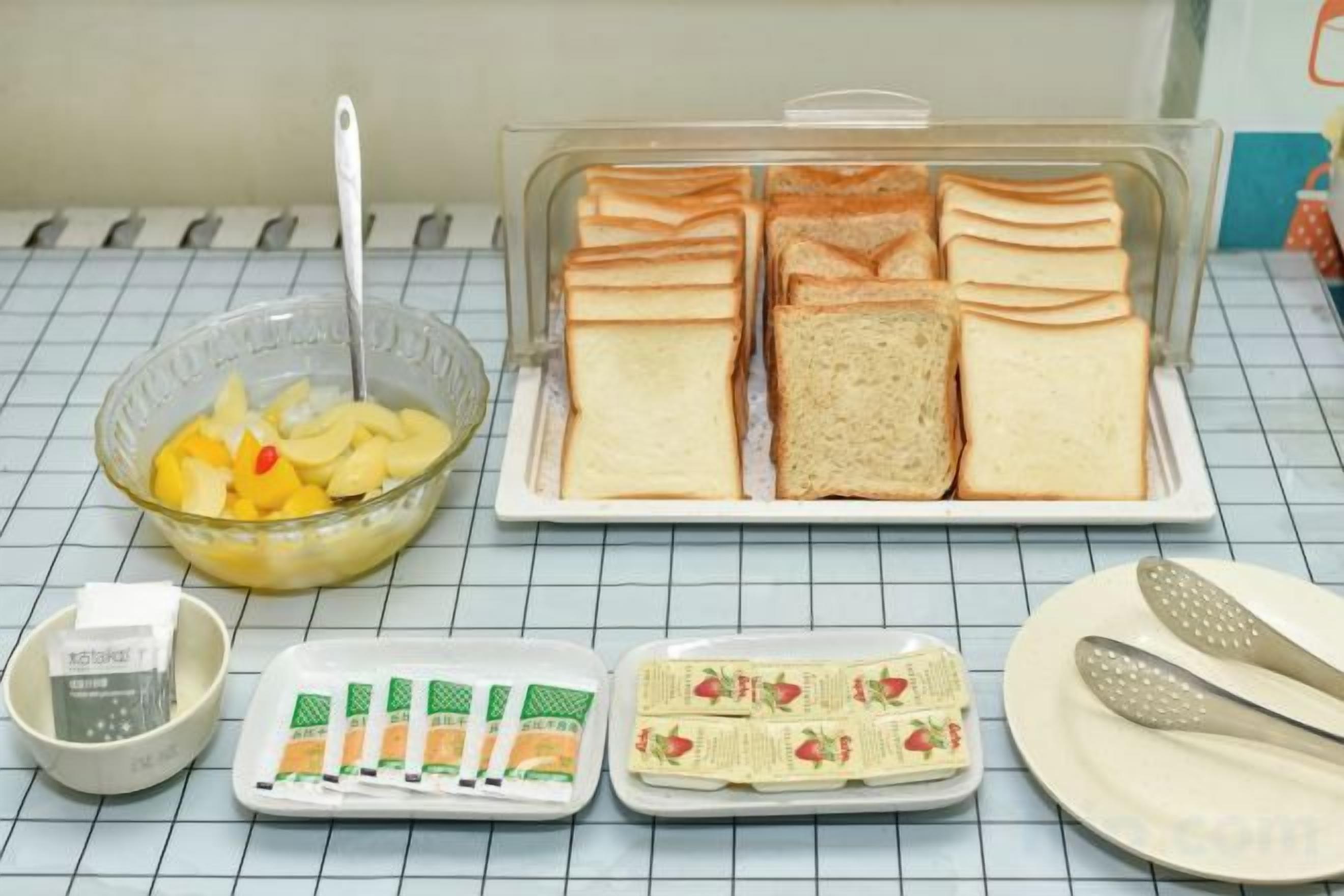 A table with a glass bowl of fruit, a plate of sandwiches, a container of condiments, and a tray of sliced bread.