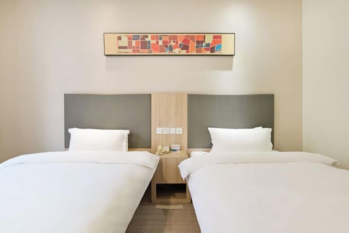 A hotel room with two single beds, a wooden headboard, a small bedside table with a phone, and a wall-mounted artwork.