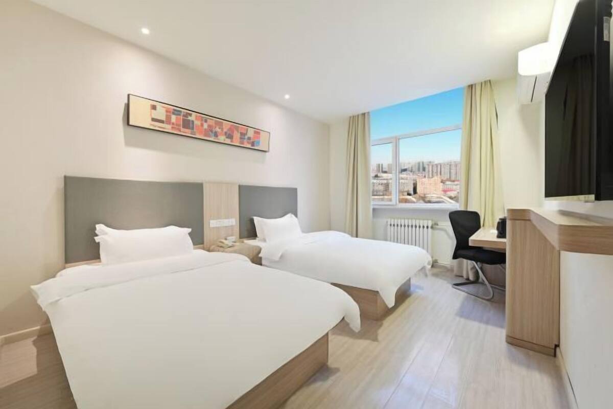 Habitación de hotel con dos camas, un escritorio, una silla, un radiador y un ventanal con vista a la ciudad.