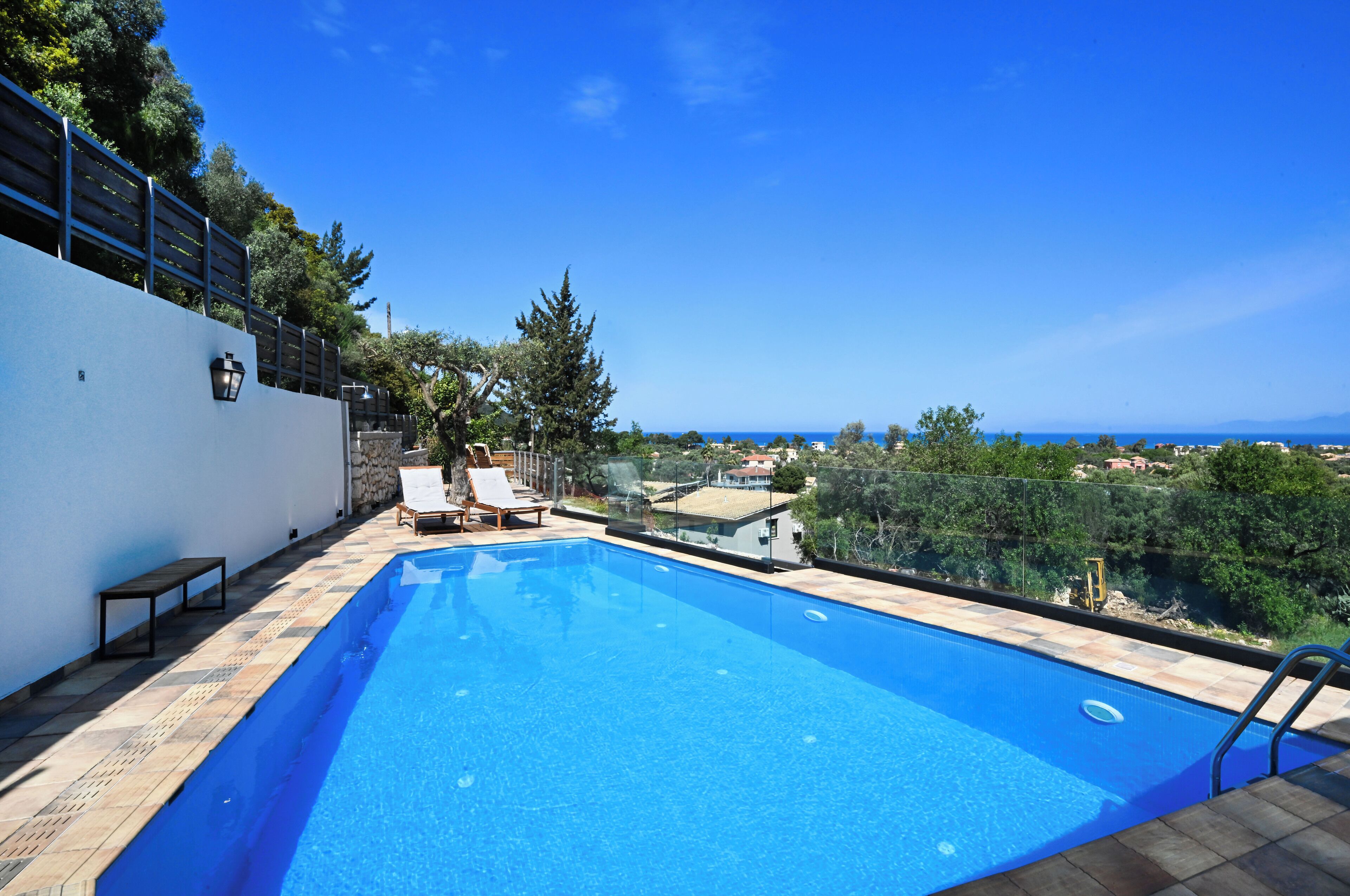 A rectangular swimming pool with a clear blue water surface, surrounded by a tiled deck. There are two lounge chairs and a small table on the deck. The pool is adjacent to a white wall with a black lamp and a glass railing. In the background, there is a view of a coastal landscape with trees and buildings.