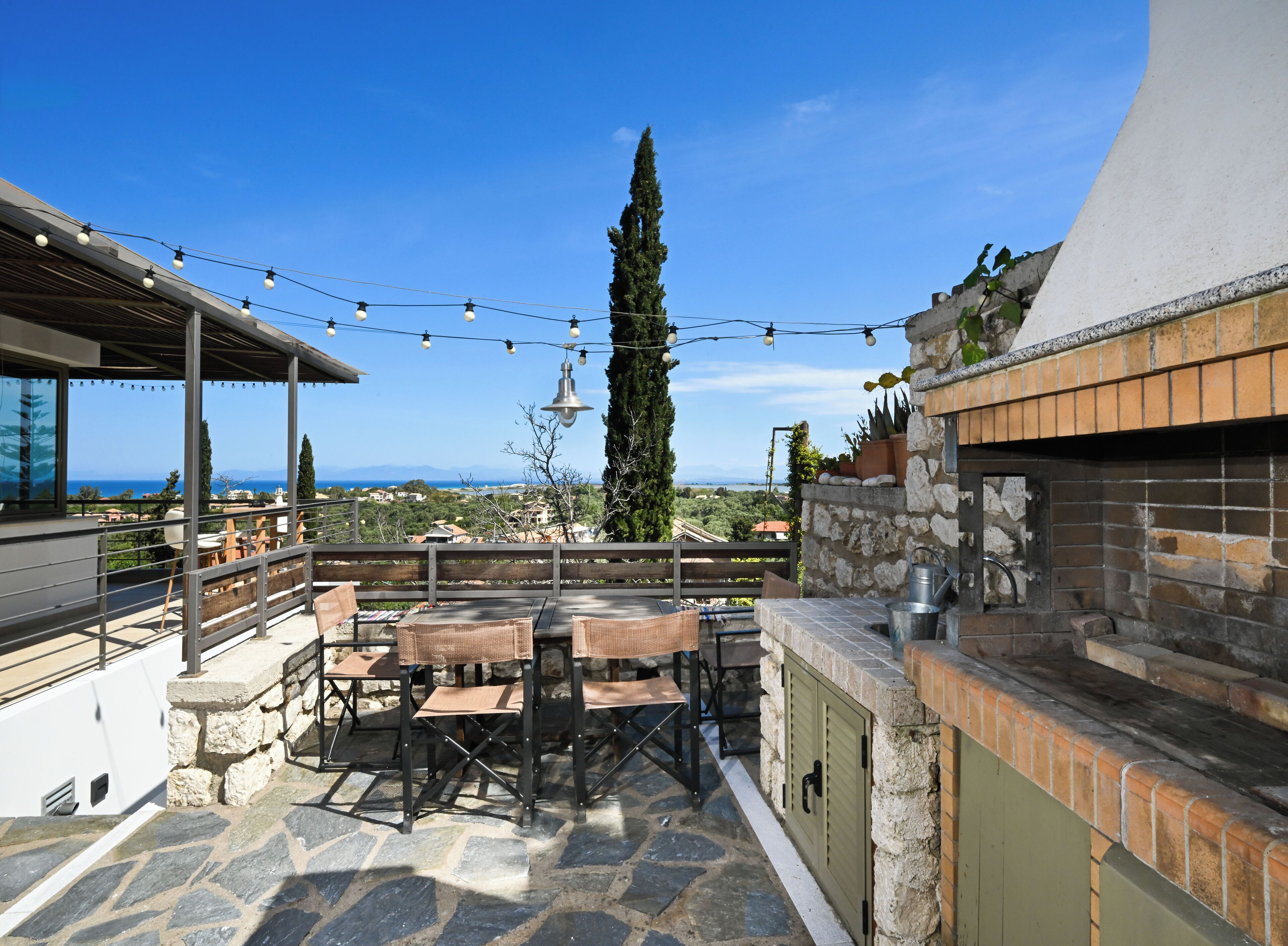 An outdoor patio with stone flooring, a wooden table and chairs, string lights, and a view of the sea and distant landscape.
