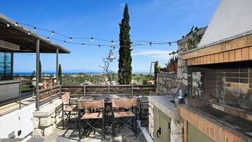 An outdoor patio with stone flooring, a wooden table and chairs, string lights, and a view of the sea and distant landscape.