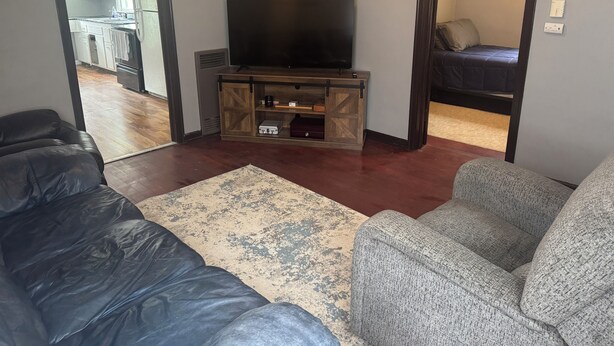 Una sala de estar con un televisor de pantalla plana sobre un soporte de madera, un sofá de cuero negro, un sillón gris y una alfombra estampada.