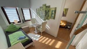 A compact bedroom with a bed, a small desk, and a chair. There are two windows with blinds, a radiator, and a staircase leading to another room. - Cozy vacation apartment - perfect for a short break all inclusive for 5 people (Bremerhaven)