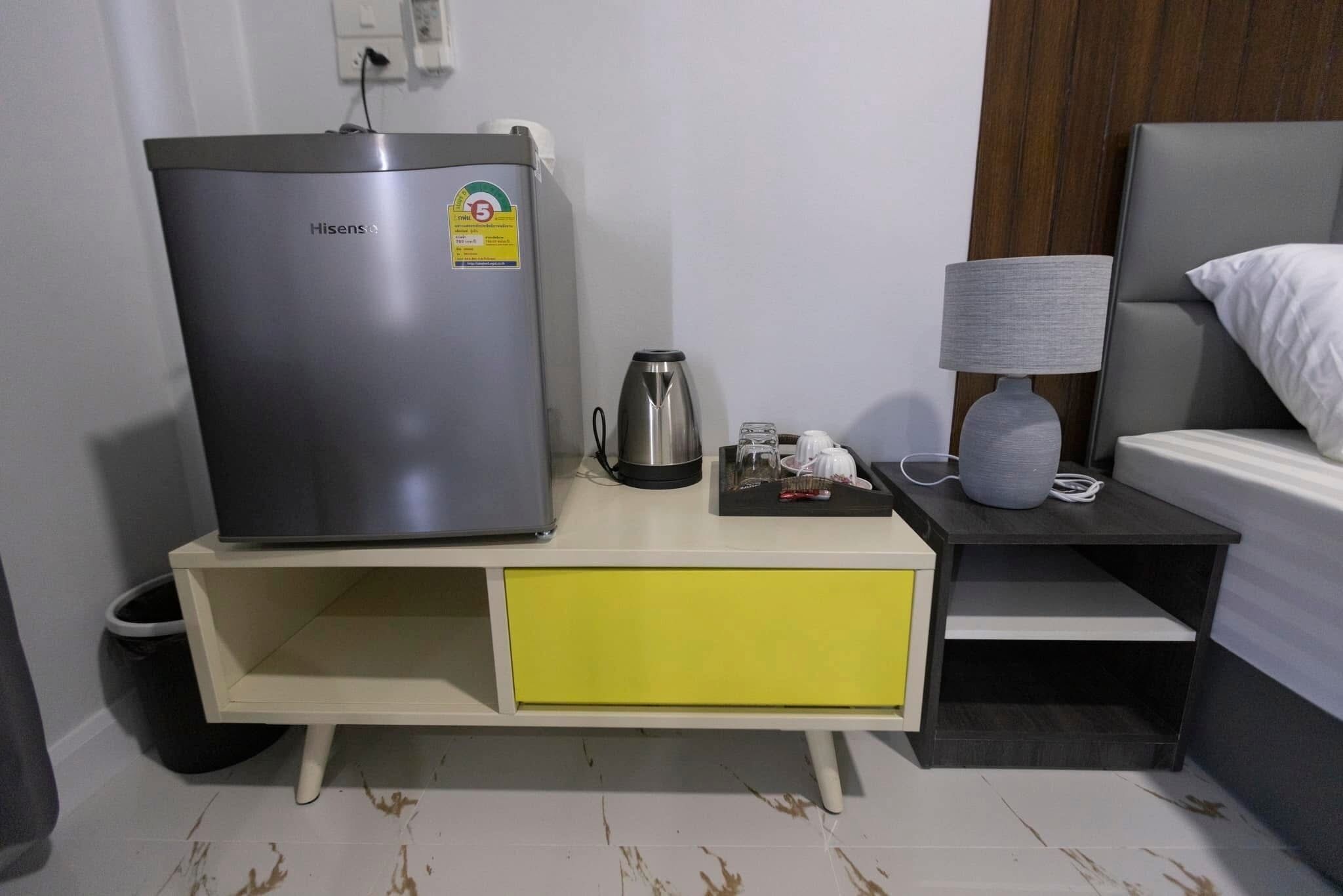 Una habitación compacta con cama, una mesita de noche con lámpara, una pequeña mesa con hervidor y un refrigerador Hisense.