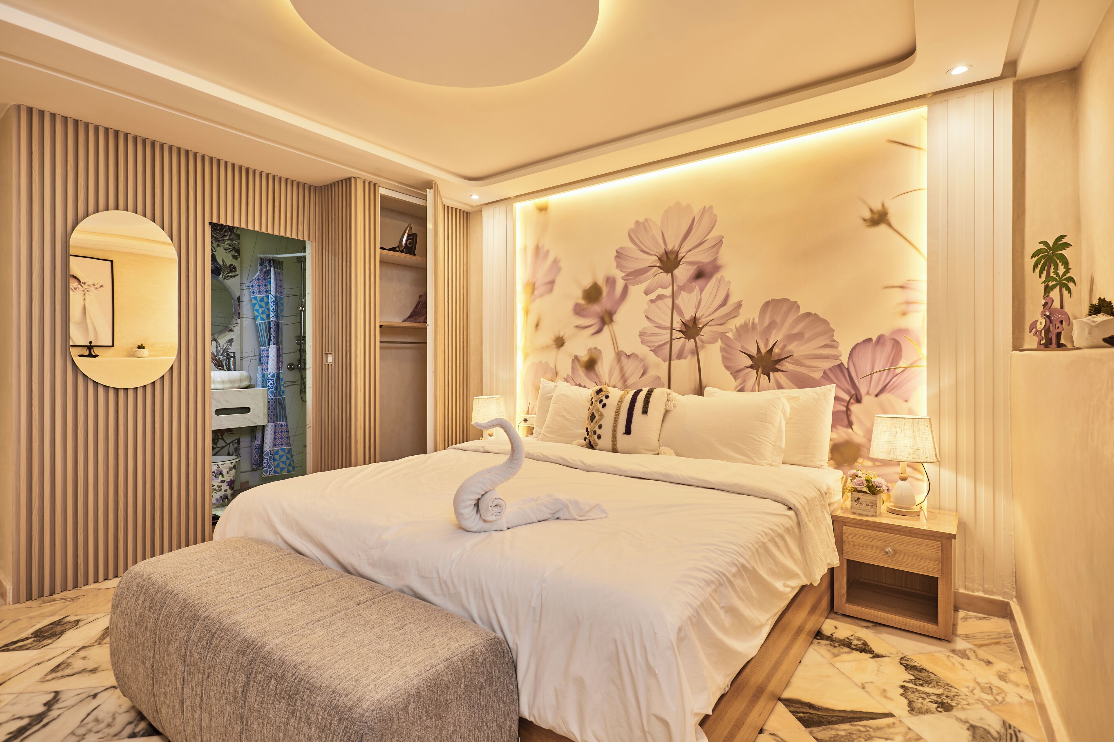 A well-lit bedroom with a large bed, a bedside table with a lamp, and a wall with floral wallpaper.
