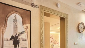 A hotel corridor with a framed picture of a historical building, a clock on the wall, and a bed visible in the adjacent room.