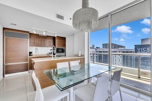 A modern kitchen with a glass dining table and white chairs, a chandelier, and a view of the city.