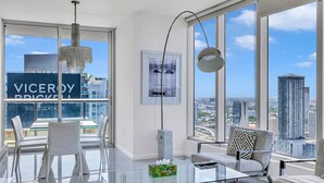 A modern living room with a glass coffee table, a sofa, and a dining area with chairs. Large windows offer a city view.