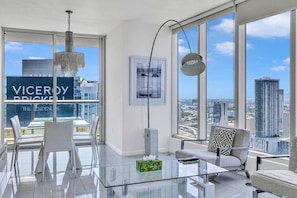 Una sala de estar moderna con una mesa de centro de vidrio, un sofá y un comedor con sillas. Amplias ventanas ofrecen vistas a la ciudad.