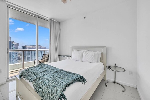 A hotel room with a bed, a small table, a chair, and a view of the city through large windows.