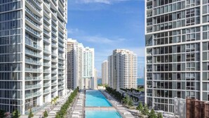 A high-rise residential building with a central swimming pool, multiple balconies, and a clear sky.