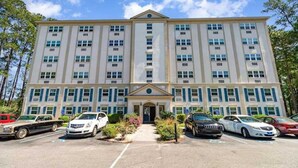A multi-story hotel with a symmetrical facade, a central entrance, and a parking lot with several cars.