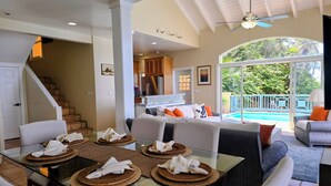 A spacious living area with a glass dining table, comfortable seating, a kitchen in the background, and a view of a swimming pool. - Ocean Front 4 Bedroom with Private Pool (St. Thomas)