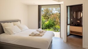 A modern bedroom with a large bed, a view of the outdoors, and a bathroom with a round mirror.