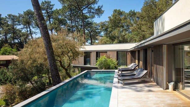 A modern outdoor pool area with a wooden deck, lounge chairs, and a view of surrounding trees.