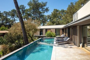 Un área de piscina moderna al aire libre con terraza de madera, sillas de descanso y vistas a árboles circundantes.