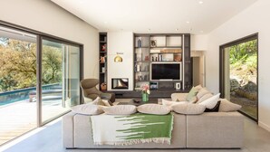 A modern living room with a large sofa, a fireplace, and a TV. There is a glass door leading to a balcony with a pool.