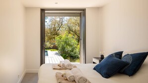 A bedroom with a large bed, a view of a garden through an open door, and a small table with a lamp.