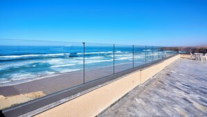A coastal terrace with a glass railing overlooking the ocean, featuring a sandy beach and a paved area with tables and chairs. - Surf & Fun Beachfront Stay in Mirleft With Balcony (Mirleft)