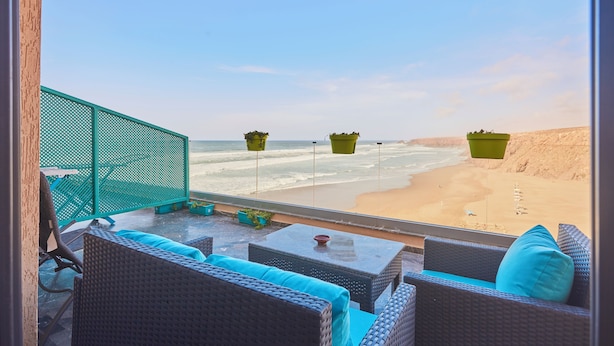 A balcony with wicker furniture, a green mesh partition, and hanging planters overlooking a sandy beach. - Surf & Fun Beachfront Stay in Mirleft With Balcony (Mirleft)