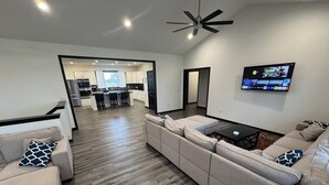 Uma sala de estar moderna com um sofá seccional, uma TV de tela plana fixada na parede e uma cozinha visível através de uma porta aberta.