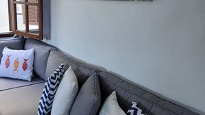 A modern living room with a grey sofa adorned with patterned pillows, a wall-mounted artwork titled "BEACH HOUSE," and a window with wooden shutters. - Beach house in Ilha Grande - Angra dos Reis - your perfect vacation on the island  (Provetá Angra dos Reis)
