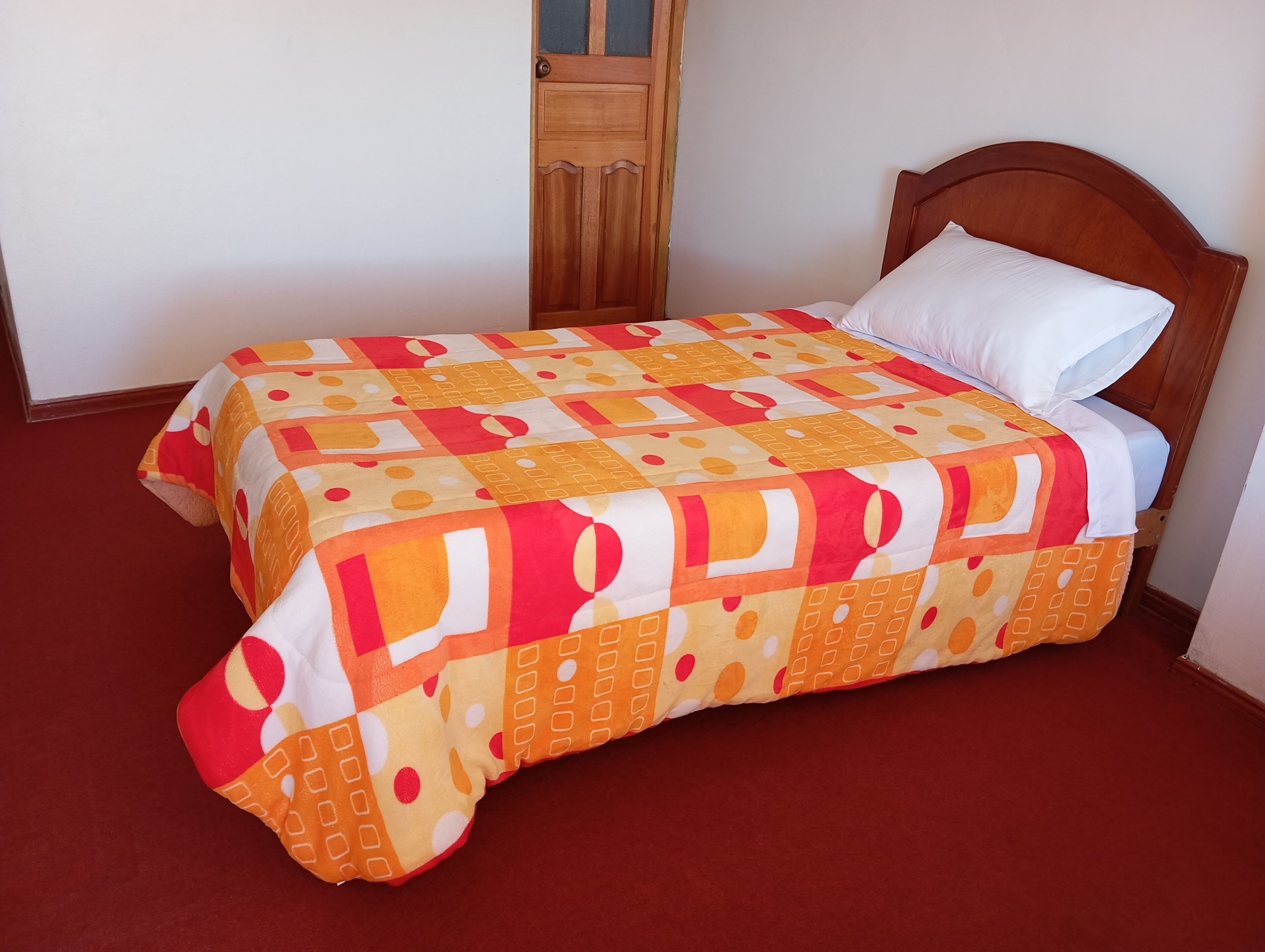 A single bed with a patterned bedspread and a white pillow in a room with a wooden door and a red carpet.