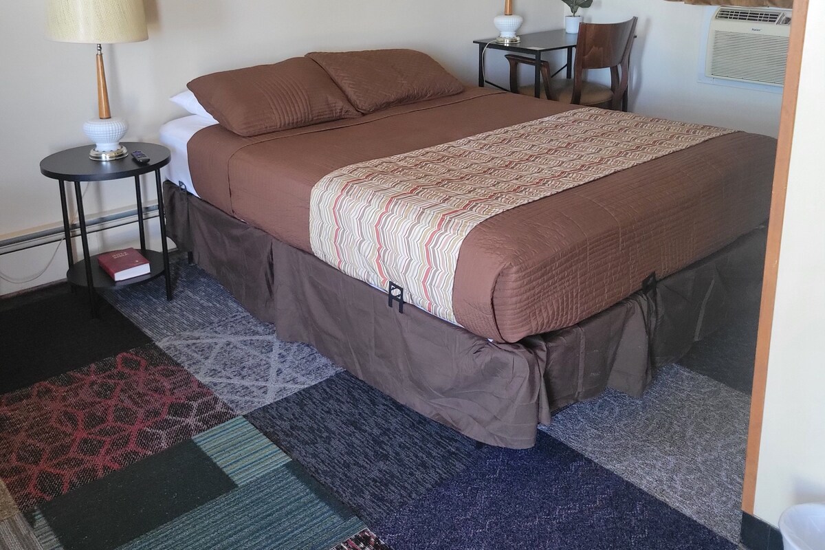 A hotel room with a large bed, two bedside tables, a lamp, a chair, a small table, a potted plant, a painting on the wall, and a window with curtains.
