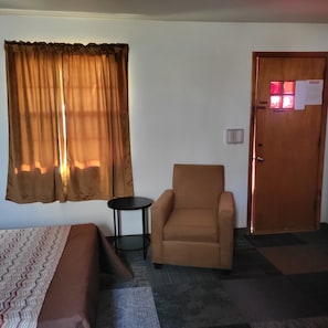 A room with a bed, a chair, a small table, a window with curtains, and a door with a 'No Smoking' sign. - Amaris Inn (Redfield)