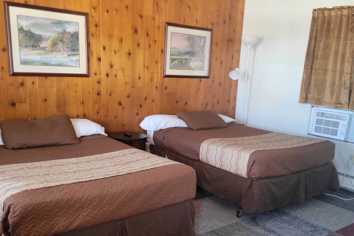 A hotel room with two beds, wooden paneling, a small table, and two framed pictures on the wall.