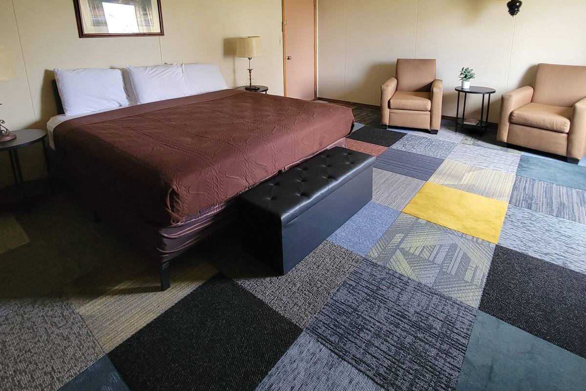 A hotel room with a bed, two chairs, a small table, a lamp, and a painting on the wall.