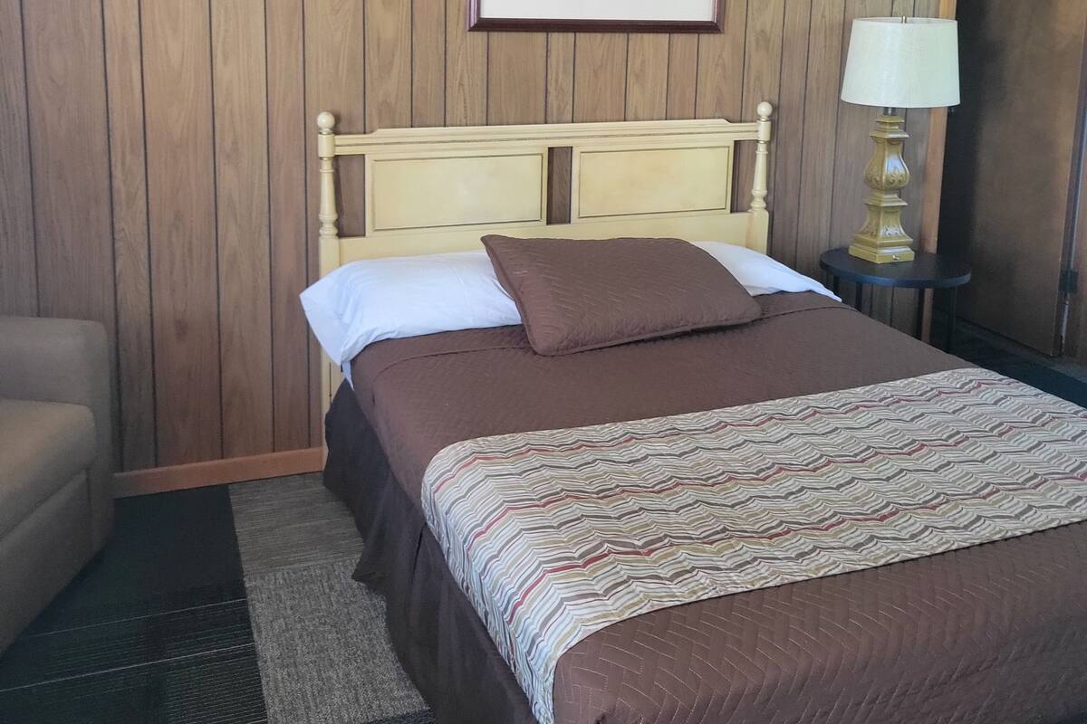 A hotel room with a wooden paneled wall, a framed landscape painting, a bed with a patterned bedspread, a bedside lamp, and a chair.