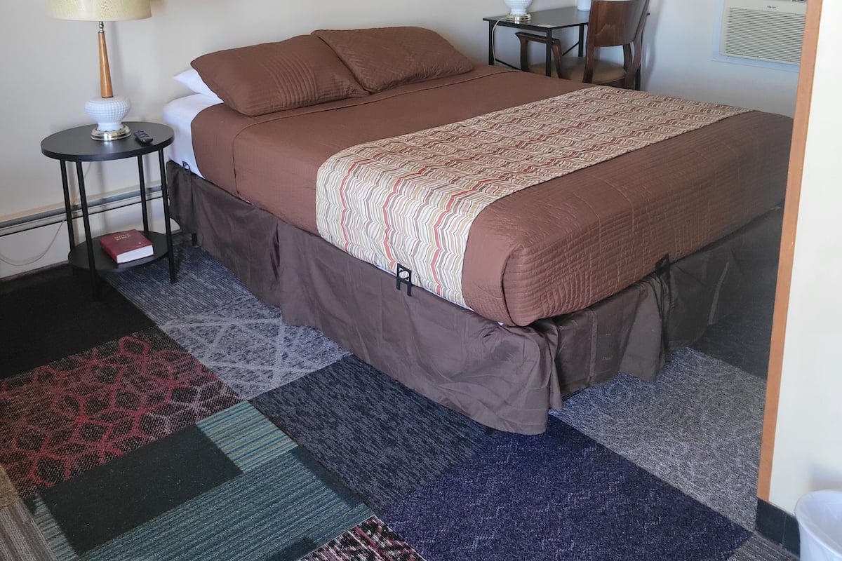 A hotel room with a large bed, two bedside tables, a lamp, a chair, a small table, a potted plant, a painting on the wall, and a window with curtains.