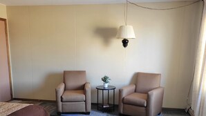 A hotel room with two armchairs, a small round table with a plant, a bed with a brown headboard, and a colorful carpet. - Amaris Inn (Redfield)