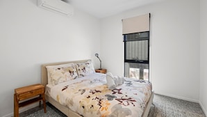 A bedroom with a bed, a nightstand, an air conditioning unit, a window with blinds, and a wall-mounted lamp. - Belmare - A Perfect Escape Surrounded by Nature (Fairhaven)
