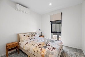 A bedroom with a bed, a nightstand, an air conditioning unit, a window with blinds, and a wall-mounted lamp. - Belmare - A Perfect Escape Surrounded by Nature (Fairhaven)