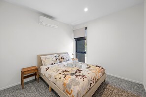 Ein Schlafzimmer mit Bett, Nachttisch, Fenster mit Jalousien und Klimaanlage.