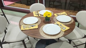 A round dining table with four white plates, yellow napkins, and silverware, surrounded by six chairs with cushioned seats. A vase with yellow flowers is placed in the center. - Del Pilar Bed & Breakfast (Managua)
