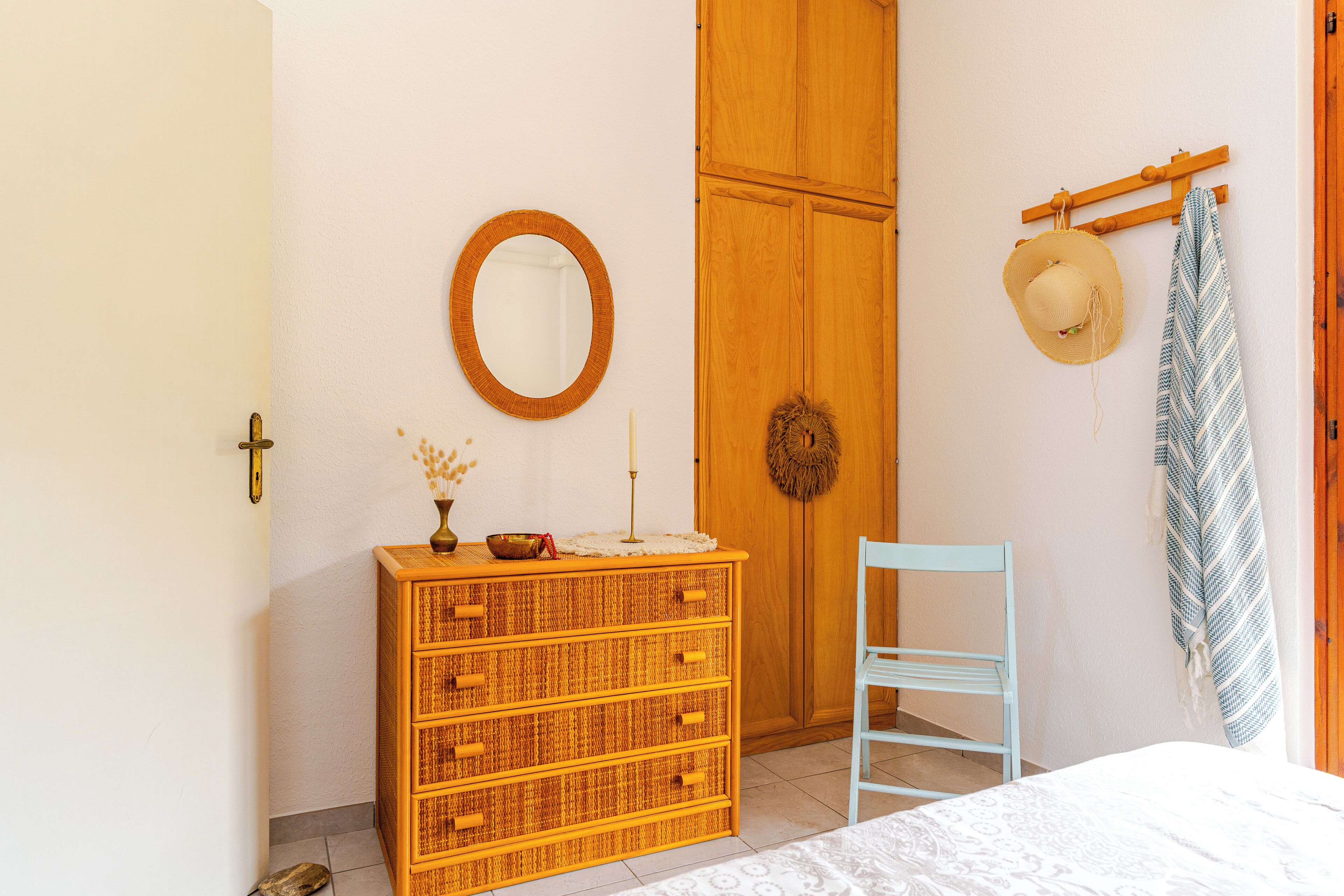 A room with a wooden wardrobe, a wicker dresser, a mirror, a hat, and a towel on a rack.