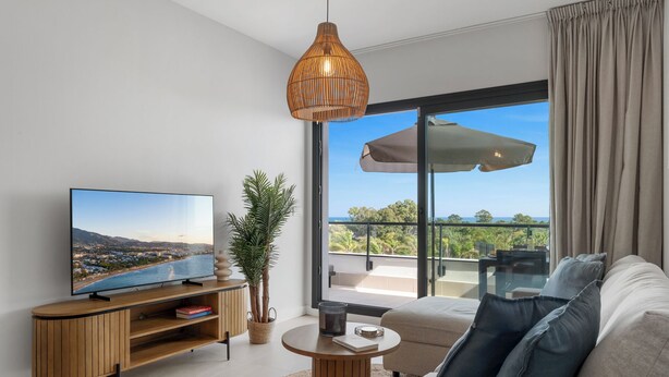 A modern living room with a flat-screen TV, a wooden entertainment unit, a round coffee table, a beige sofa, a potted plant, and a large sliding glass door leading to a balcony with a view of the sea. - Top Floor Seaview Apartment (Estepona)
