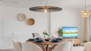 Un comedor moderno con una mesa redonda, sillas color beige y un televisor mostrando una escena de playa.