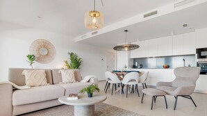 Una sala de estar moderna con un sofá, una mesa de centro y sillones, un comedor con una mesa y sillas, y una cocina con armarios y electrodomésticos blancos.