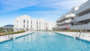 Una piscina con superficie de agua azul cristalina, rodeada de edificios blancos con balcones, y un cielo despejado arriba.