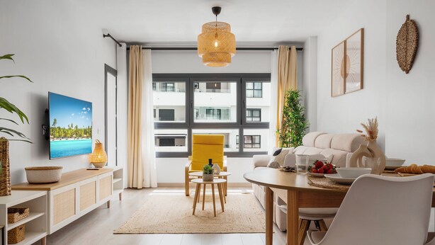 Una sala de estar moderna con un sofá, una mesa de comedor y sillas. Hay un televisor en la pared, una ventana con cortinas y una planta.