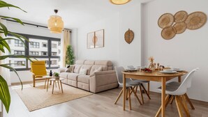 Una sala de estar moderna con un sofá color beige, una mesa de comedor con sillas, una butaca amarilla y un ventanal con vistas a edificios.