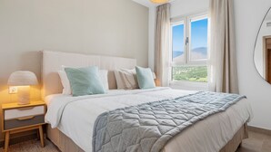 A bedroom with a bed, a nightstand, a lamp, a window with a view, and a circular mirror on the wall.
