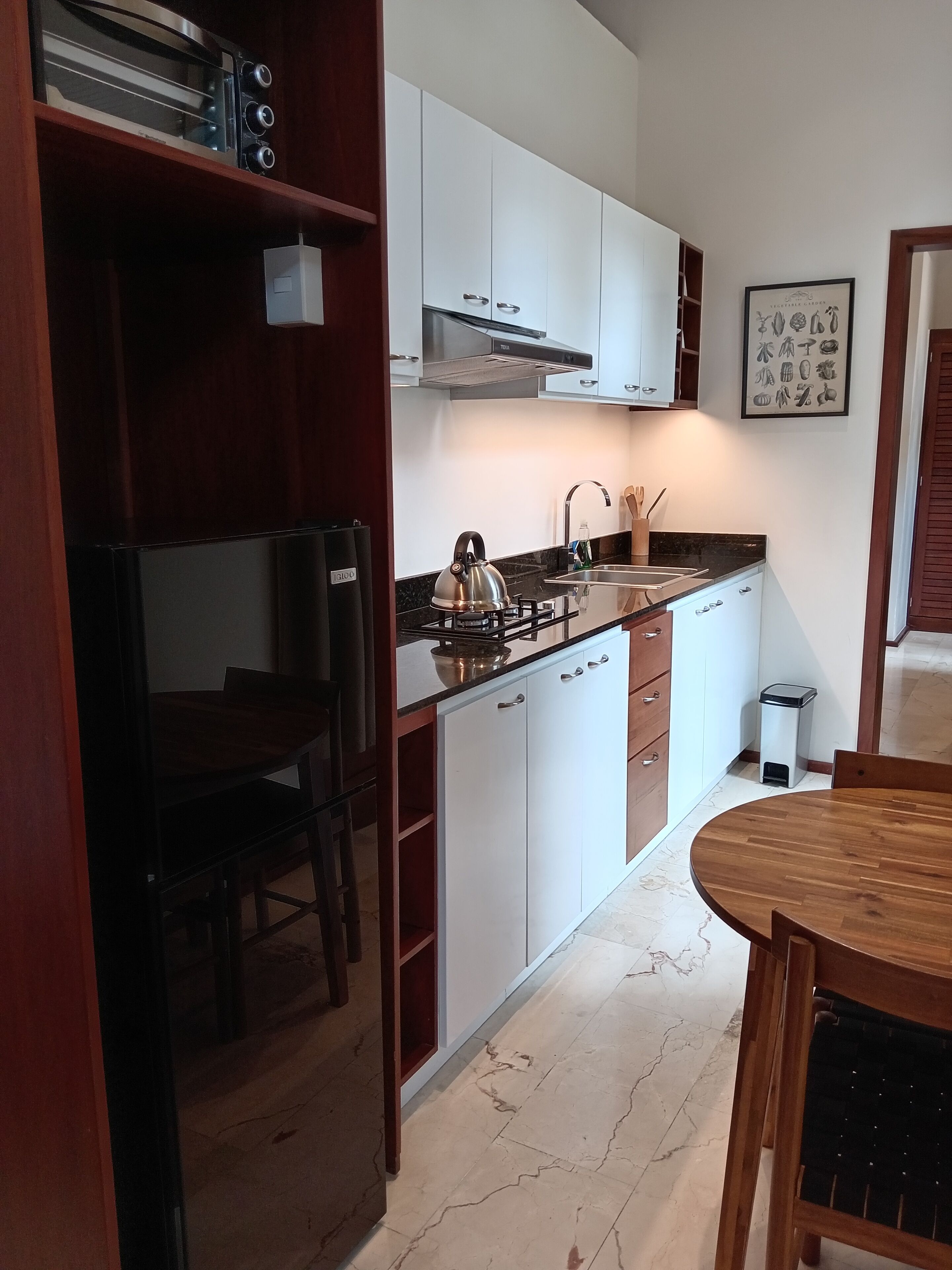 Una cocina moderna con armarios blancos, un hervidor de acero inoxidable y una estufa a gas. Se observa una mesa de comedor de madera y sillas, además de un cubo de basura.
