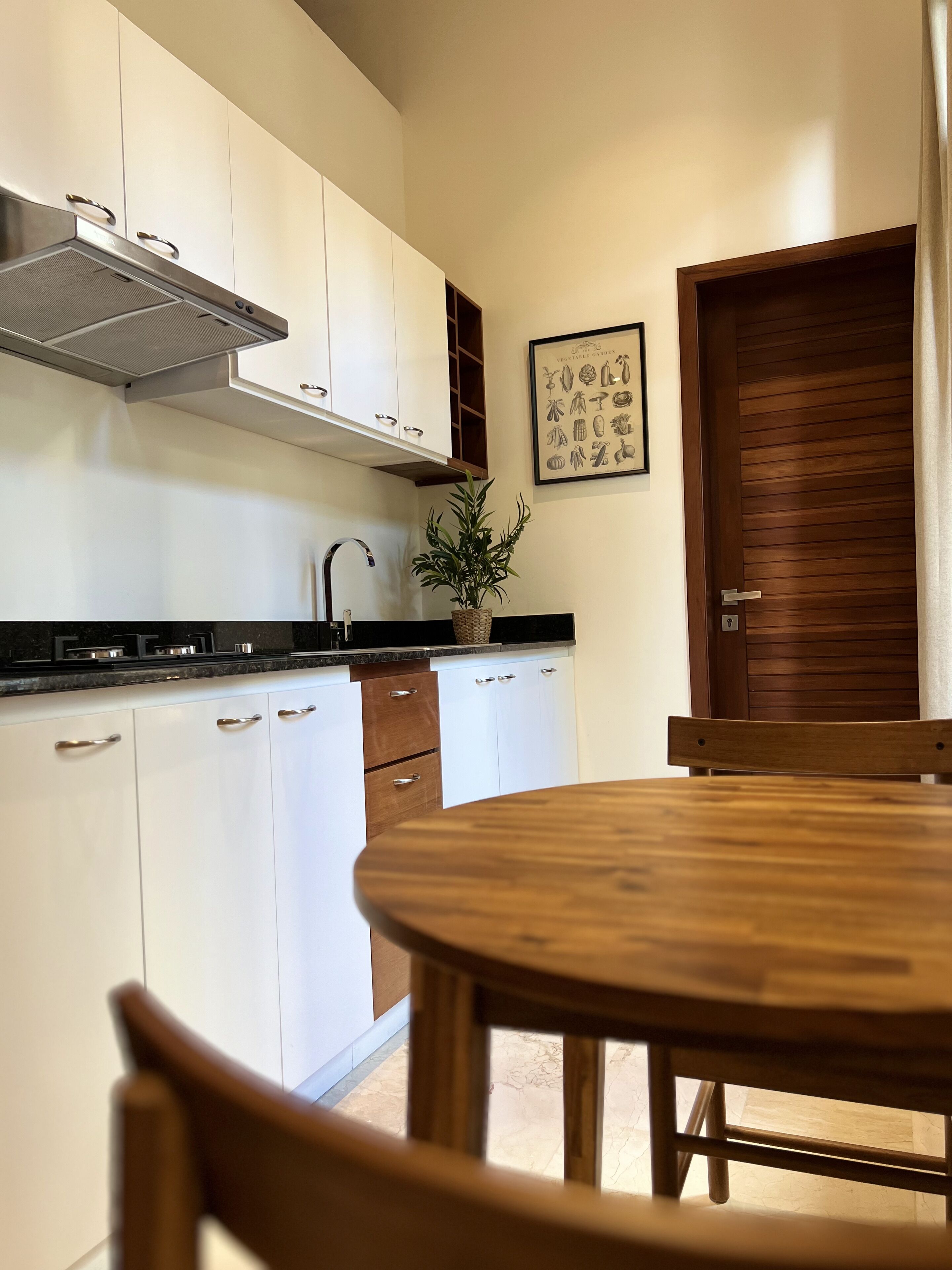 Una cocina con armarios blancos, una campana extractora de acero inoxidable, una mesa de comedor de madera y una planta en maceta.