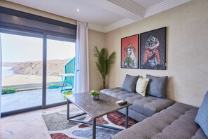 Relax in this stylish living room with a sofa, coffee table, and artwork, leading to a balcony with a view of the beach and rocky terrain.